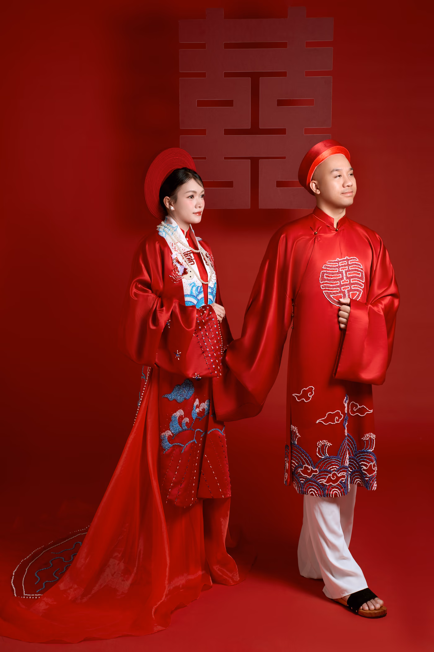 Cô dâu và chú rể trong áo dài đỏ thêu hoa văn mây sóng, đội khăn đóng đỏ, đứng cạnh nhau trước tấm phông đỏ có chữ song hỷ.