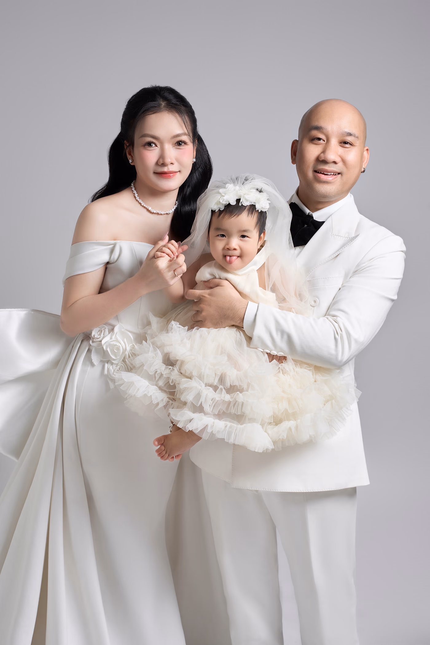 Cô dâu bế một bé gái mặc váy voan trắng, chú rể vest trắng thắt nơ đen đứng cạnh ôm cả hai, nền xám nhạt.