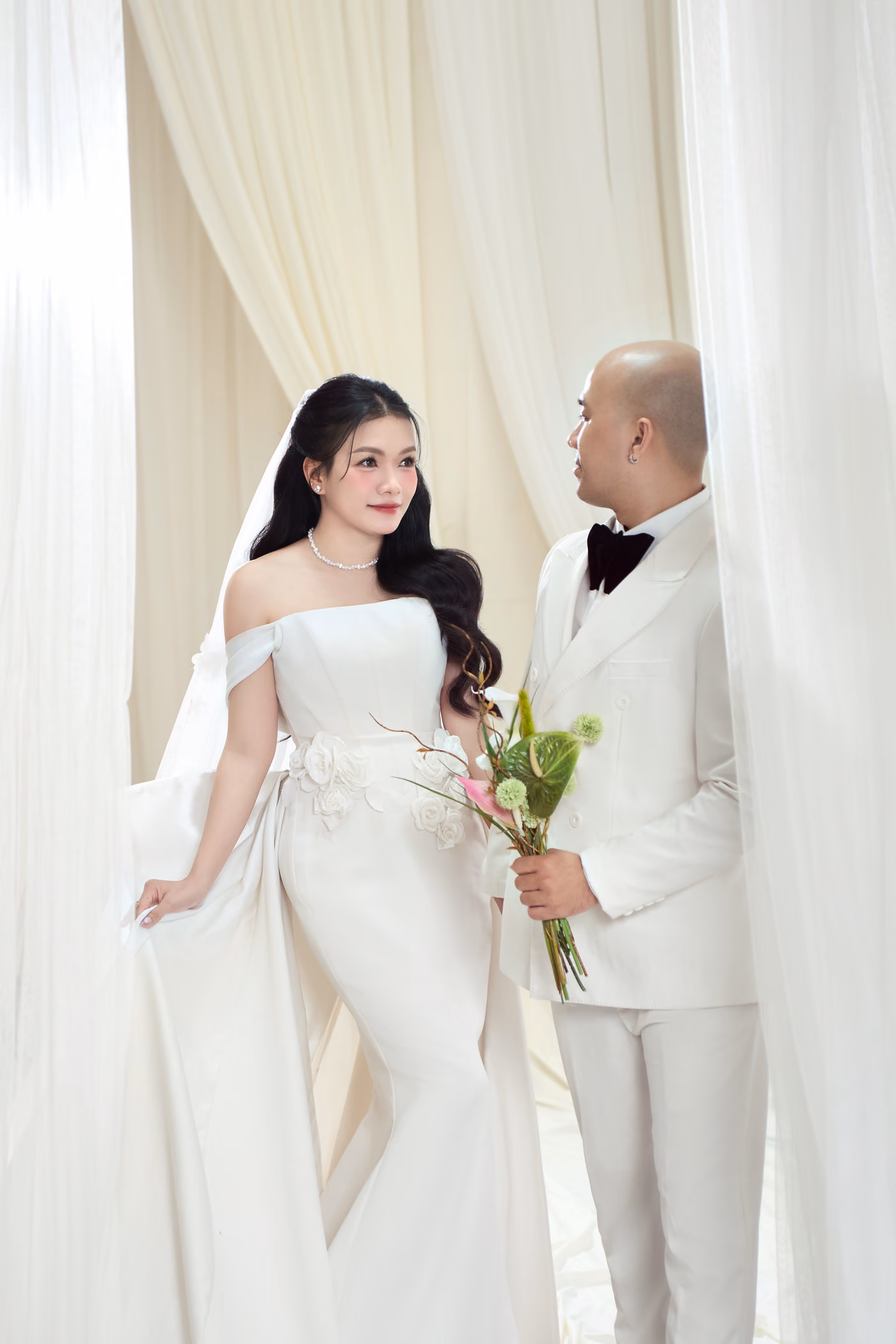 Cô dâu váy trắng ôm dáng và chú rể vest trắng cầm bó hoa nhỏ đứng giữa những tấm rèm voan màu kem.