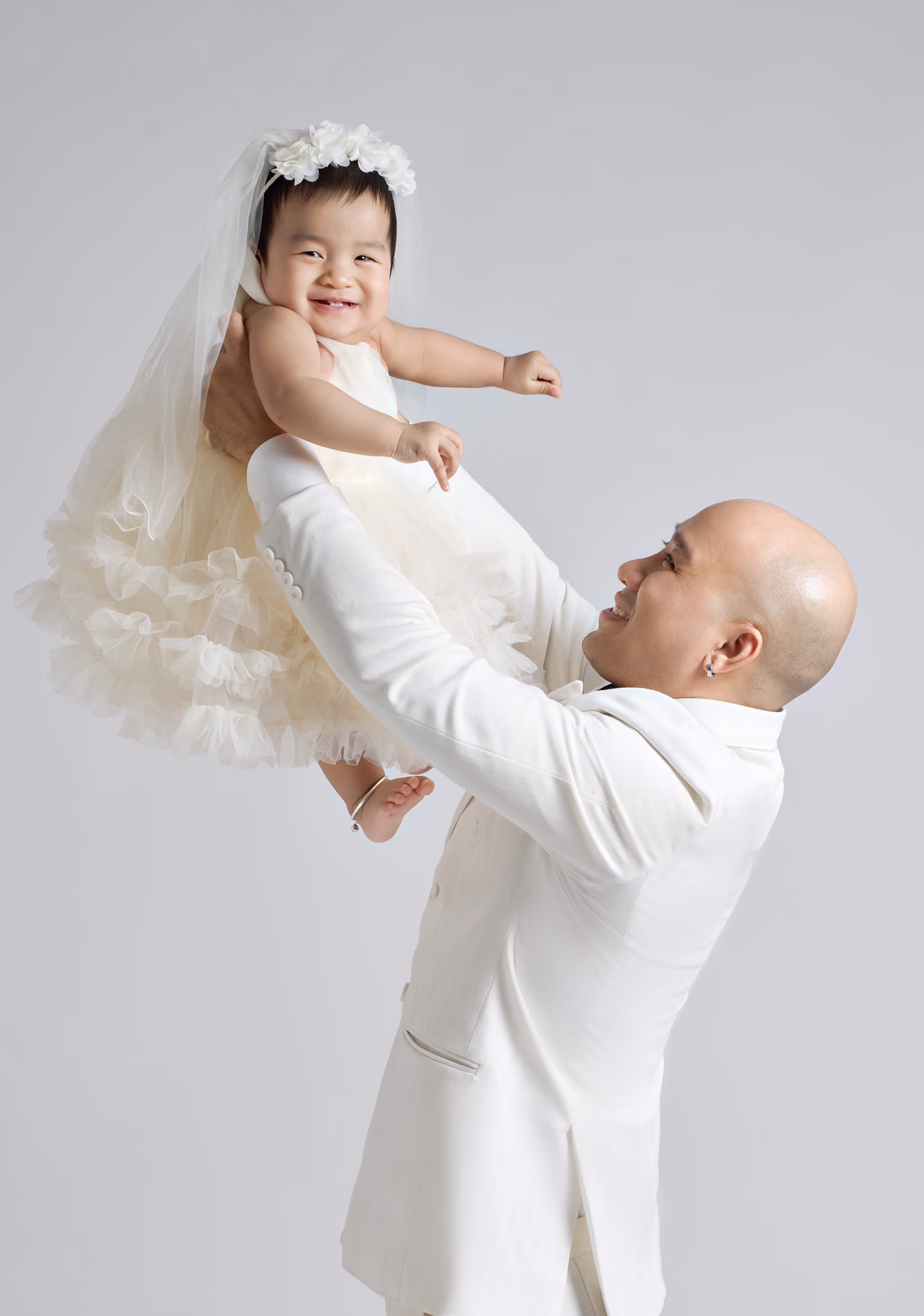 Chú rể vest trắng nâng bé gái mặc váy voan bồng và đội vương miện hoa lên cao, cả hai cười, nền xám nhạt.