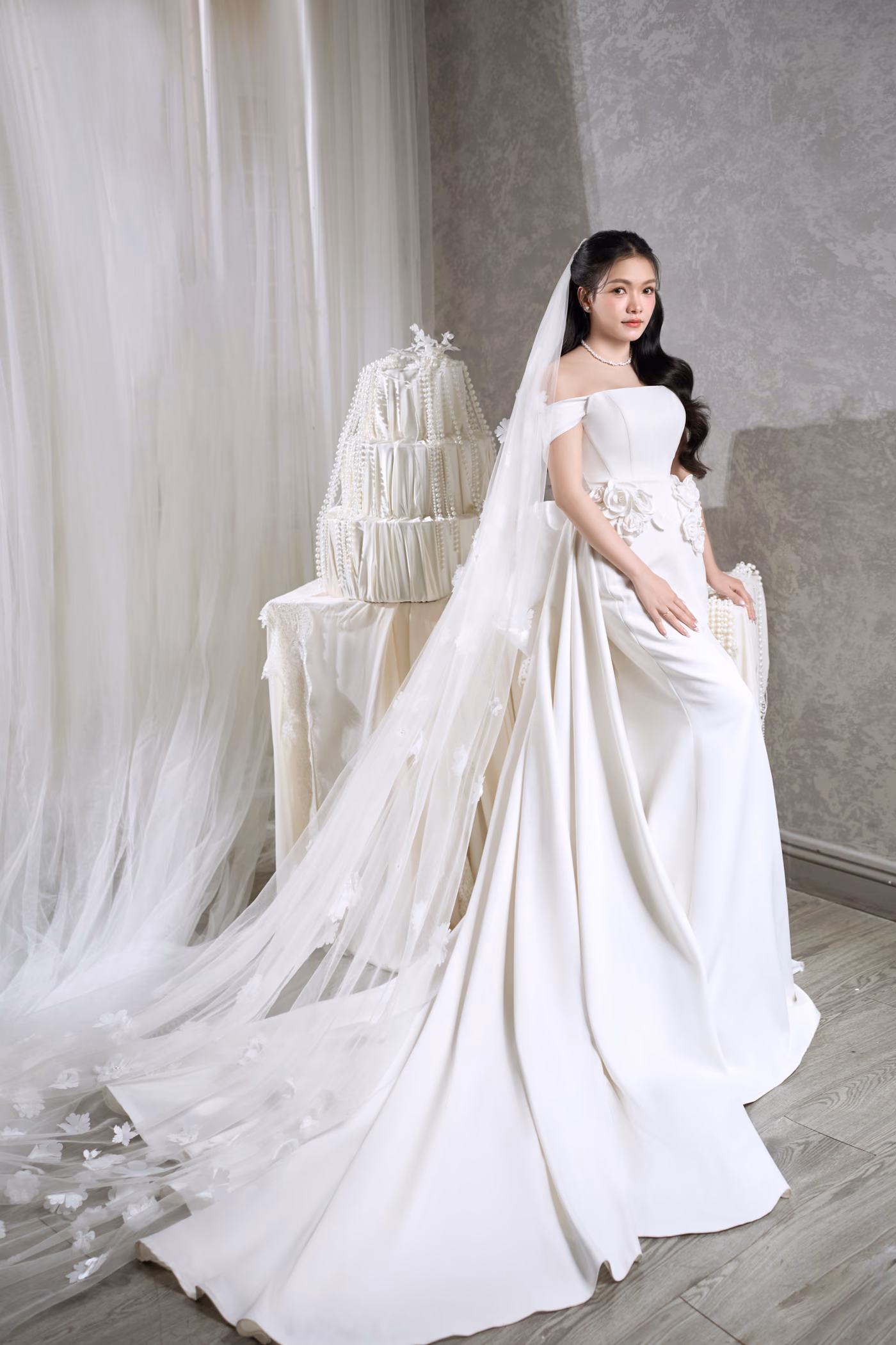 Cô dâu váy cưới trắng với tấm voan dài buông trên sàn gỗ, đứng cạnh chiếc bánh cưới nhiều tầng giữa những tấm rèm trắng.