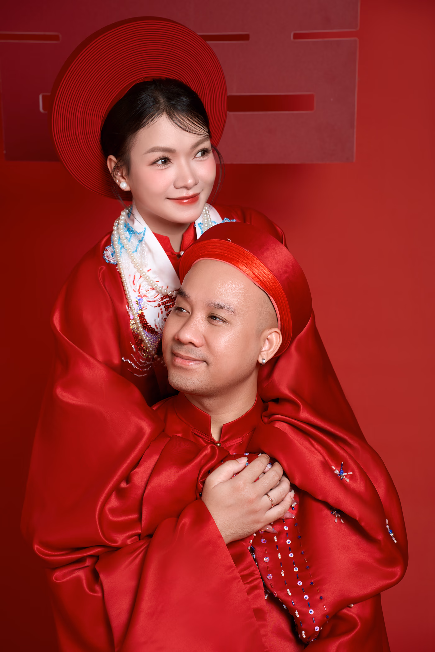 Cận cảnh cô dâu và chú rể kề sát nhau trong áo dài đỏ và khăn đóng đỏ, trước nền đỏ có chữ song hỷ.