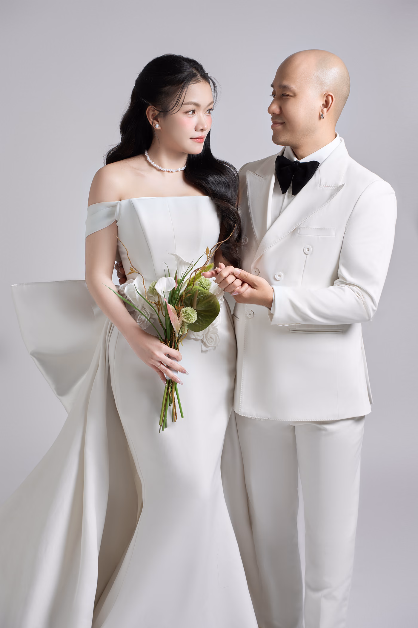 Cô dâu váy trắng trễ vai cầm bó hoa nhỏ và chú rể vest trắng thắt nơ đen nắm tay nhau, nhìn nhau mỉm cười trên nền xám nhạt.