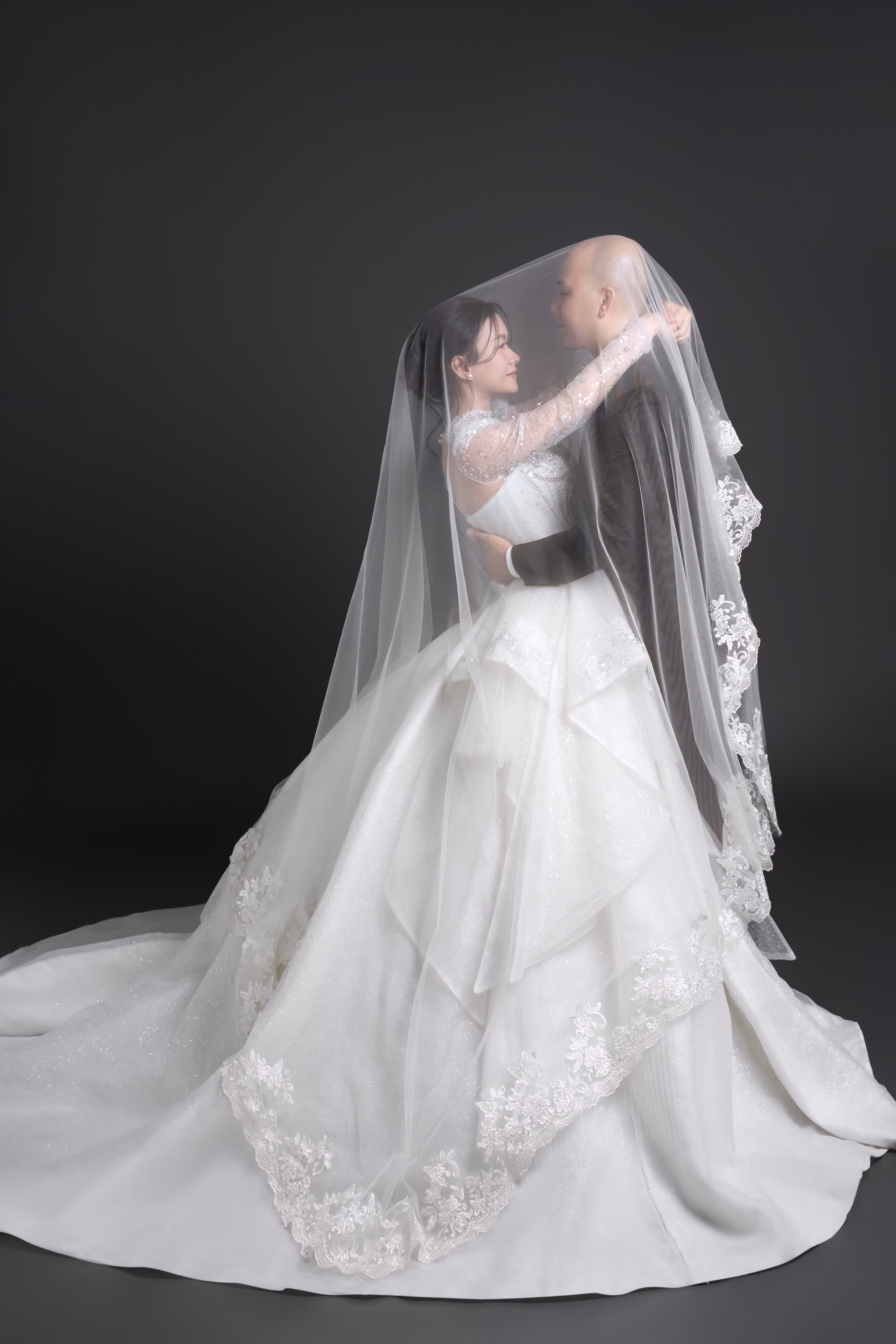 Cô dâu và chú rể ôm nhau dưới tấm voan dài viền ren; cô dâu mặc váy cưới xếp tầng, nền xám than.