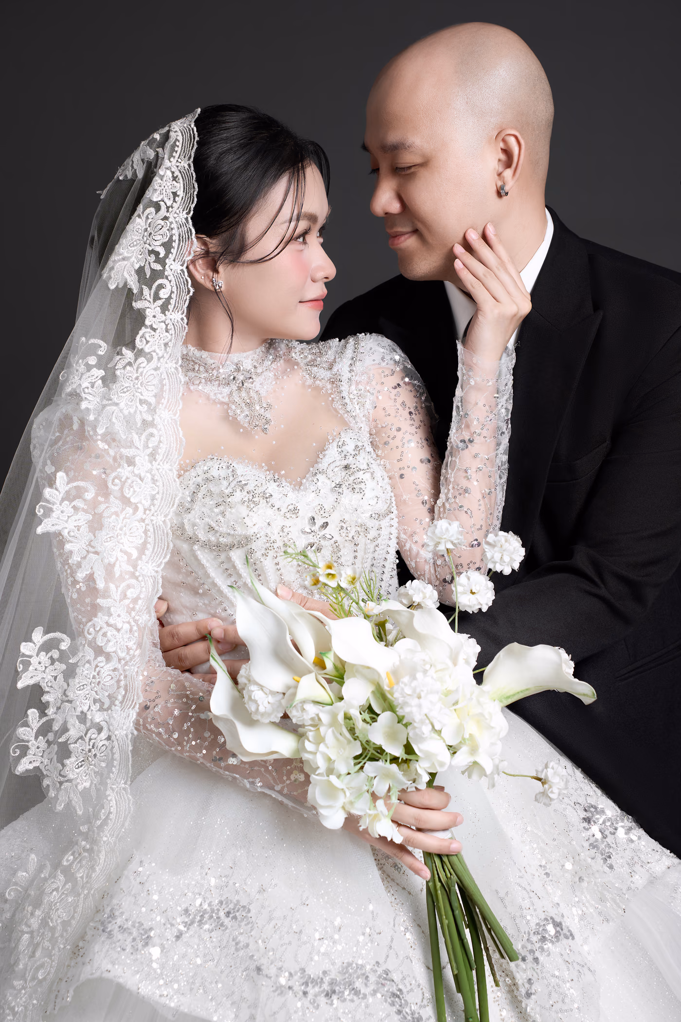 Cận cảnh cô dâu đội voan ren, váy đính hạt, cầm bó hoa calla trắng, một tay chạm nhẹ vào cằm chú rể mặc vest đen đang nghiêng đầu về phía cô.