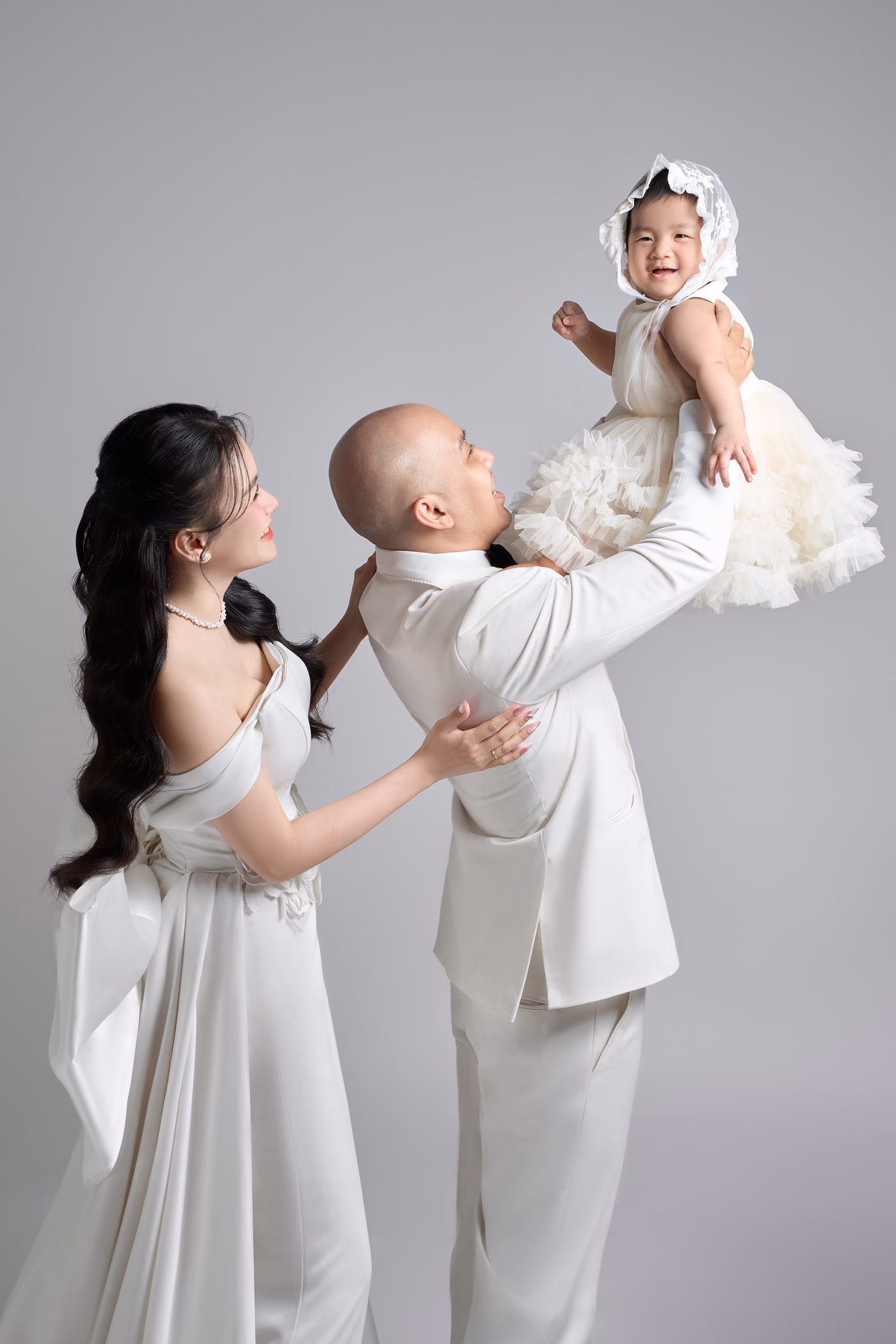 Chú rể vest trắng nâng một bé gái mặc váy voan bồng và đội mũ ren lên cao, cô dâu váy trắng trễ vai đứng bên cạnh đỡ bé, nền xám nhạt.